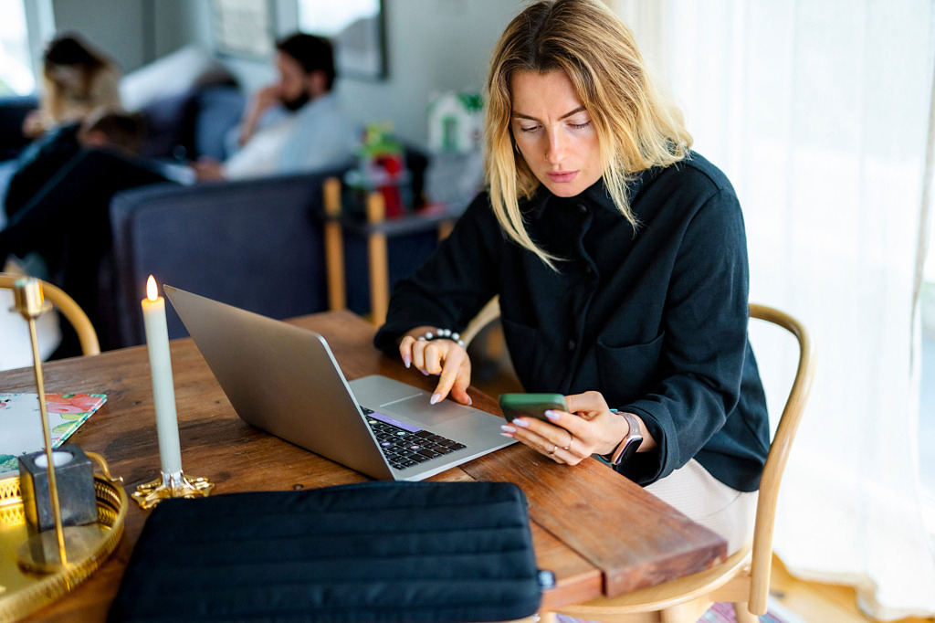 Kvinna som sitter vid ett matbord och använder telefon och laptop.