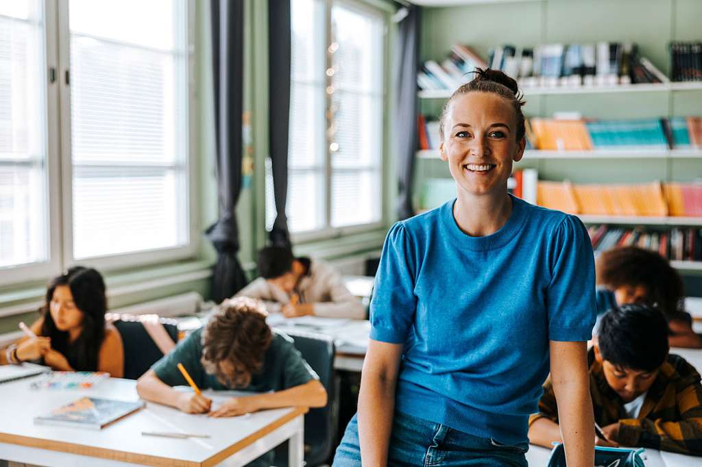 Lärare sitter på en bänk i ett klassrum med elever i bakgrunden som skriver i skrivblock.