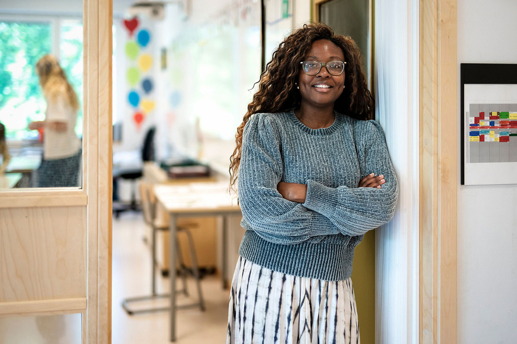 Lärare står med armarna i kors i öppningen till klassrummet. Hon ser glad ut.